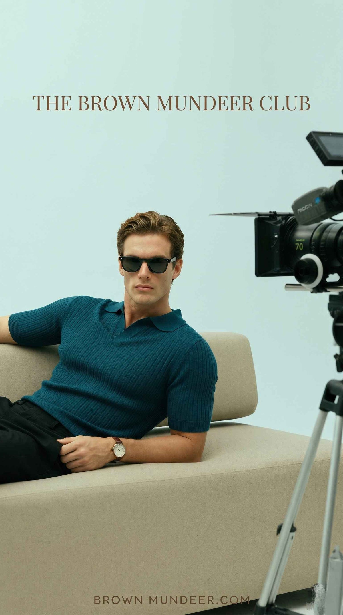 Man in a ribbed teal polo shirt and sunglasses lounging on a beige sofa during a photoshoot.